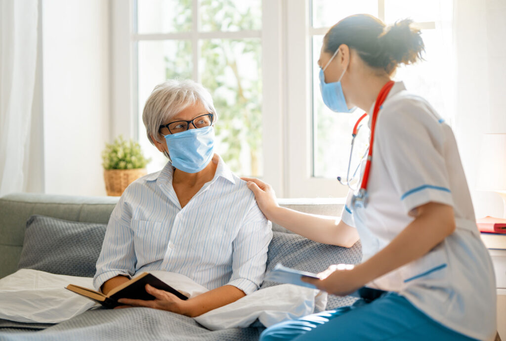 female nurse speaking with senior wearing face masks on a couch