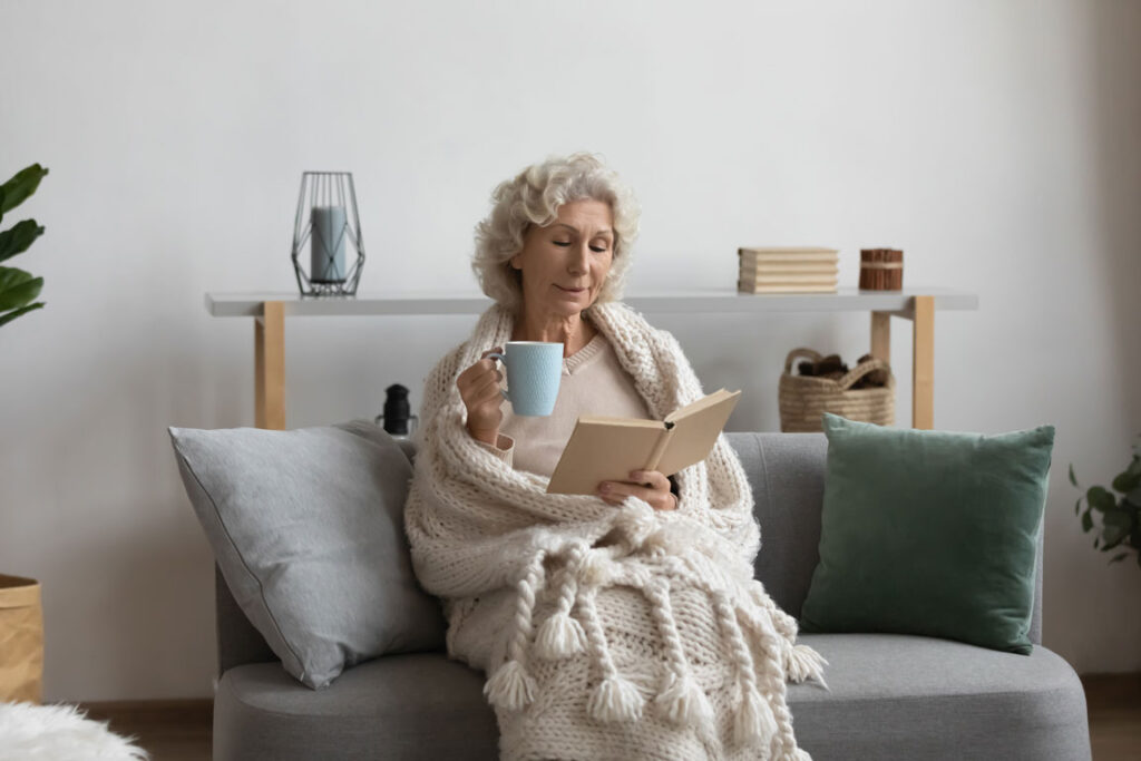 senior woman on couch drinking coffee and reading a book wrapped in a blanket