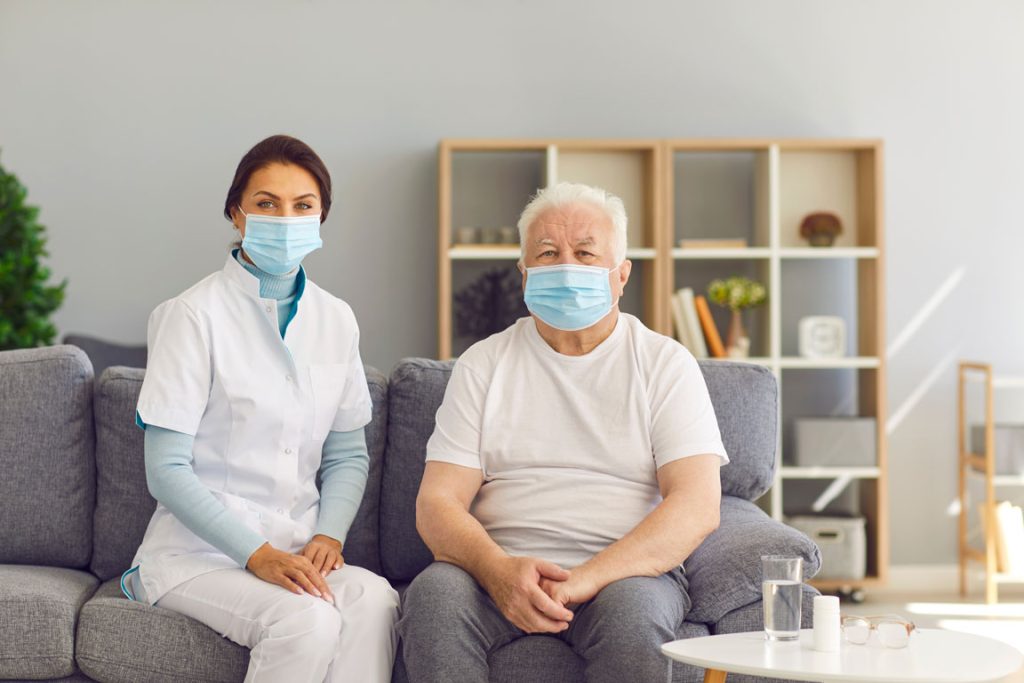 nurse and nursing home resident sitting on sofa wearing protective face masks